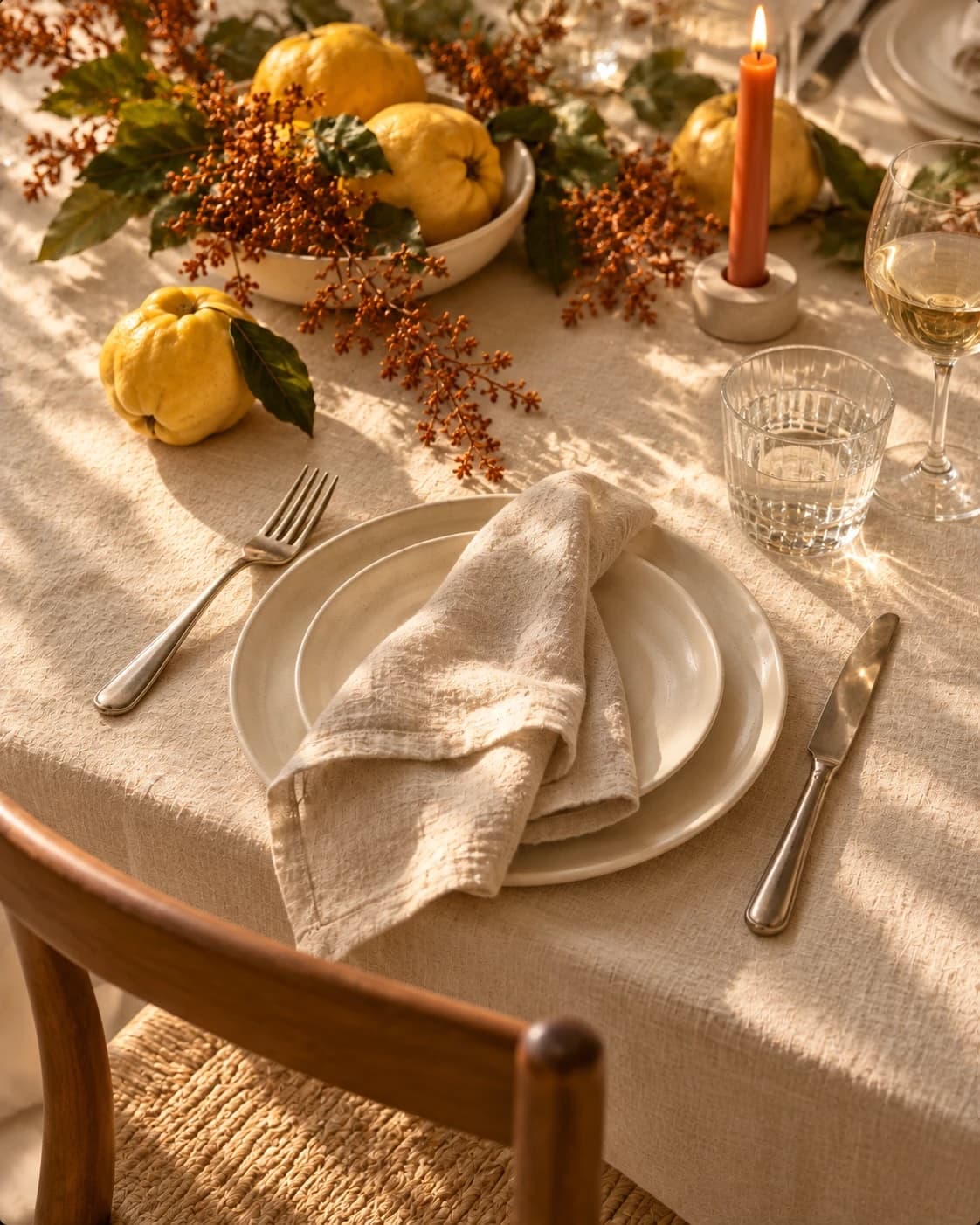 A place setting with a linen napkin on stoneware plates, a quince and autumn branches beyond, by a wooden chair.