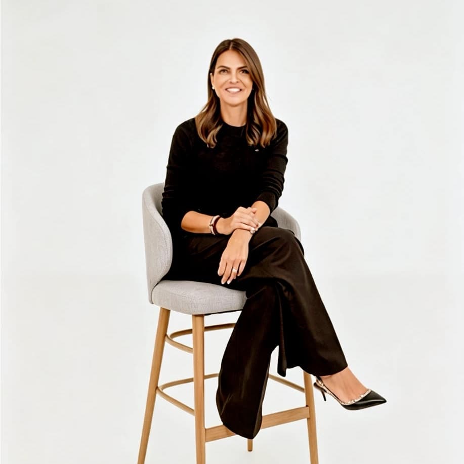 Zahra Abdalla smiling, seated on a tall stool in a black jumper and trousers against a white backdrop.