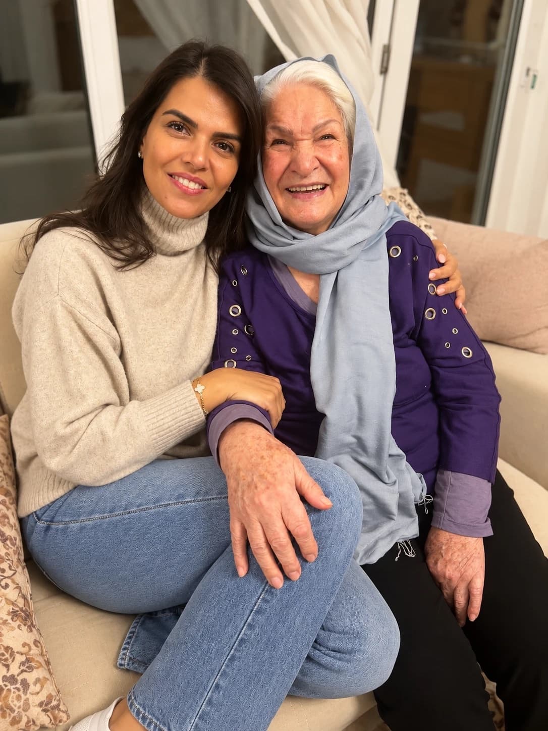 Two women smiling, sitting close together on a cream sofa, one in a beige jumper and the other in a pale blue headscarf and purple top.
