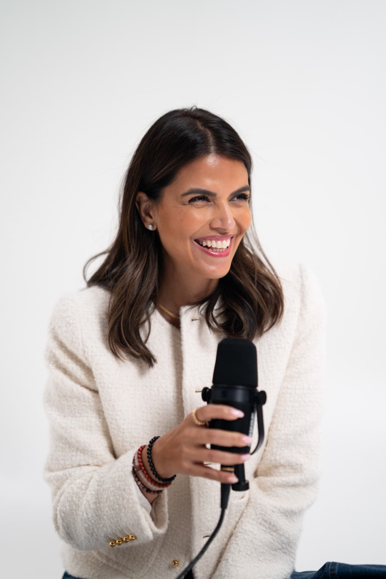 Zahra Abdalla laughing as she holds a podcast microphone, in a cream bouclé jacket against a white backdrop.